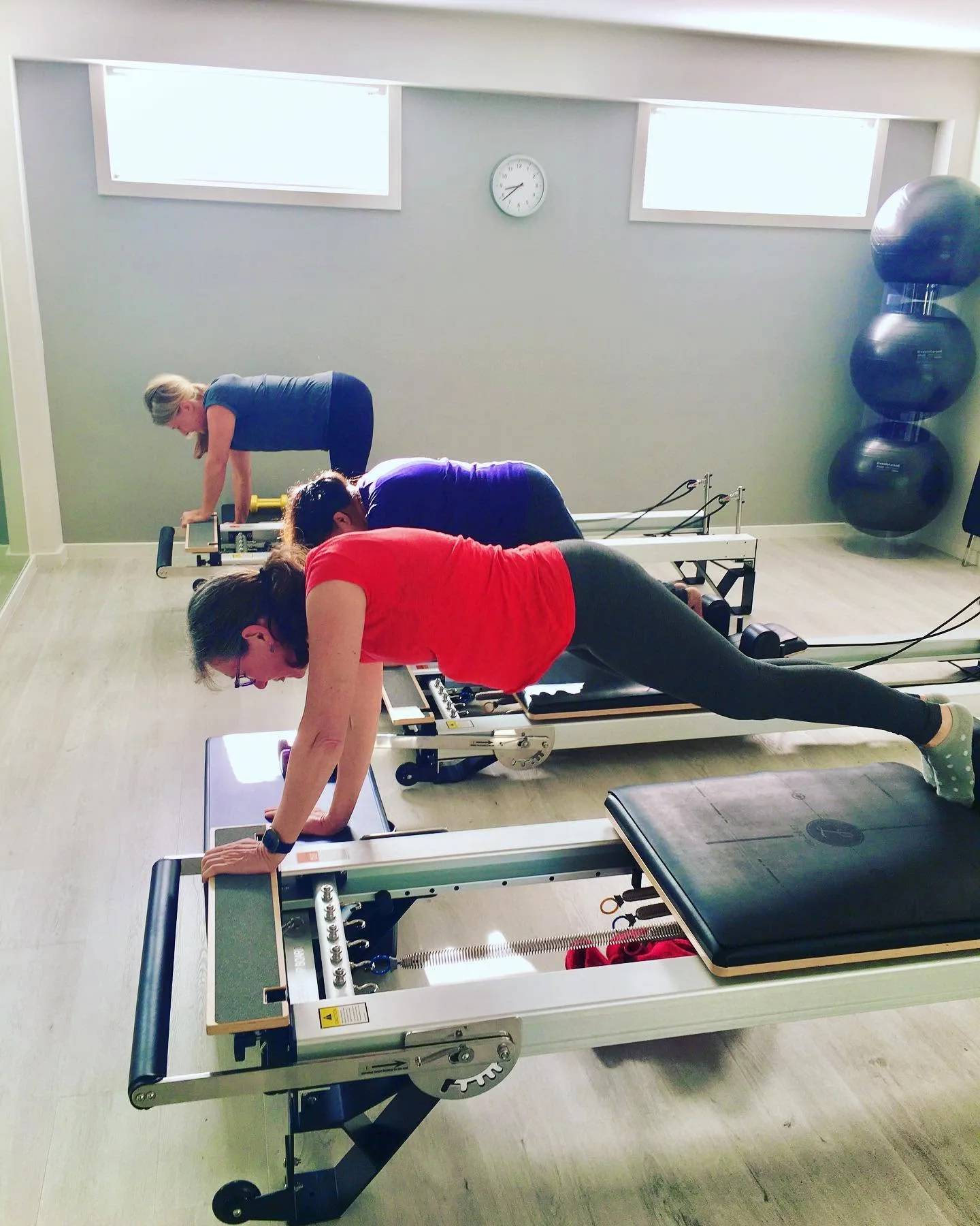 Pilates class holding plank on reformers, stability balls and wall clock in view